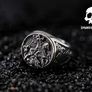 May include: A silver ring featuring an intricate design of a knight on horseback. The ring is displayed against a black backdrop.