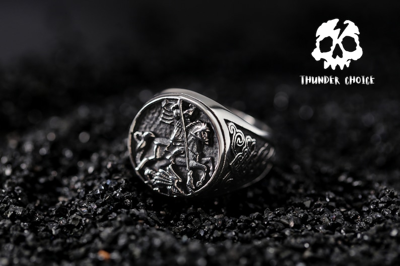 May include: A silver ring featuring an intricate design of a knight on horseback. The ring is displayed against a black backdrop.