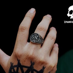 May include: A silver ring featuring a detailed design of two figures on horseback. The ring is worn on the index finger of a hand with a black tattoo.