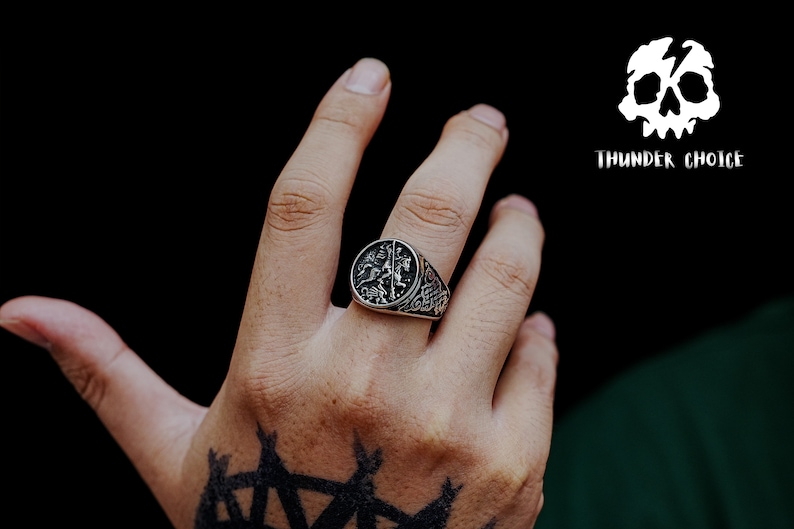 May include: A silver ring featuring a detailed design of two figures on horseback. The ring is worn on the index finger of a hand with a black tattoo.