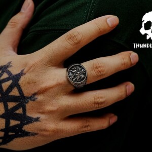 May include: A silver ring with an intricate design featuring a cross and other symbols. The ring is worn on the index finger of a hand with a black tattoo.