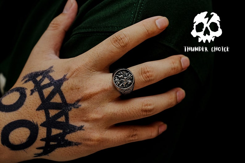 May include: A silver ring with an intricate design featuring a cross and other symbols. The ring is worn on the index finger of a hand with a black tattoo.