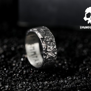 Peut inclure: Une bague en argent avec une surface texturée et rugueuse. La bague est sur un fond noir.