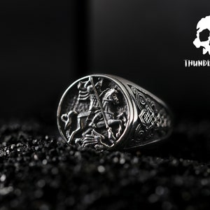 May include: A silver ring featuring an intricate design of a knight on horseback slaying a dragon. The ring is displayed against a black backdrop.