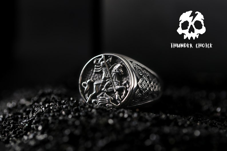 May include: A silver ring featuring an intricate design of a knight on horseback slaying a dragon. The ring is displayed against a black backdrop.