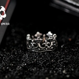 May include: A silver crown ring with intricate details. The ring is on a black background.