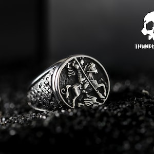 May include: A silver ring featuring an intricate design of a knight on horseback. The ring is set against a black backdrop.