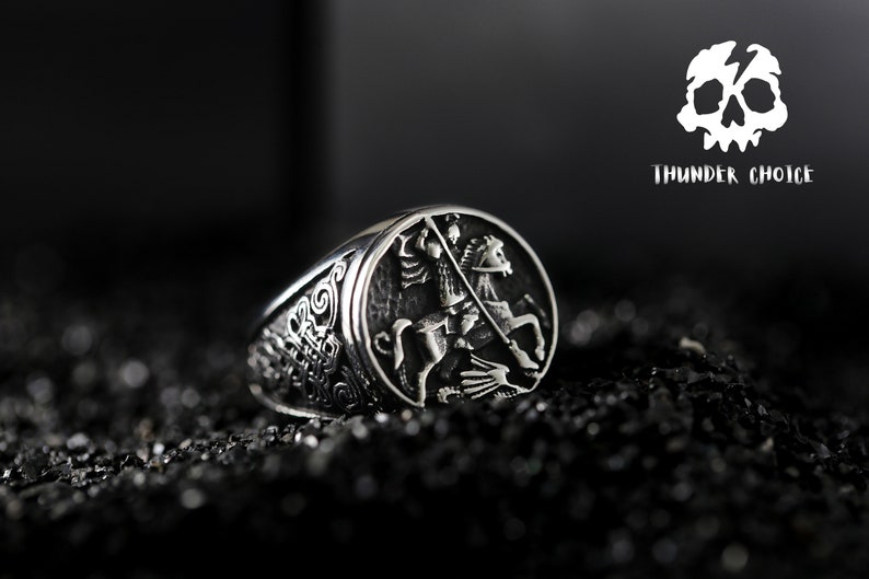 May include: A silver ring featuring an intricate design of a knight on horseback. The ring is set against a black backdrop.