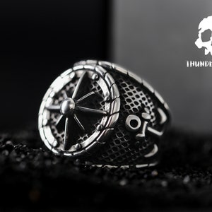 May include: A silver ring featuring a compass design and a mesh pattern on the band. The ring is set against a black background.