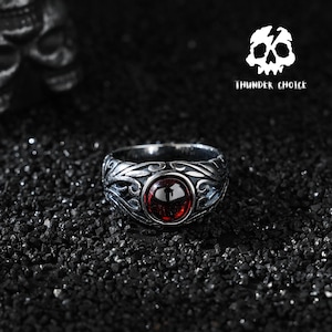 May include: A silver-toned metal ring featuring an intricate, raised vine-like pattern and a central, smooth, circular red gemstone. The ring is positioned near a logo depicting a white skull with the text THUNDER CHOICE underneath it.