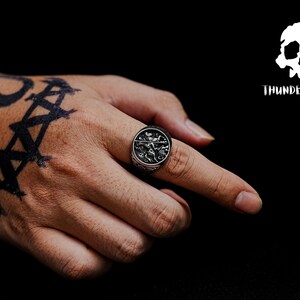 May include: A silver ring with a detailed design featuring a skull and crossbones. The ring is worn on the index finger of a hand with a black tattoo.