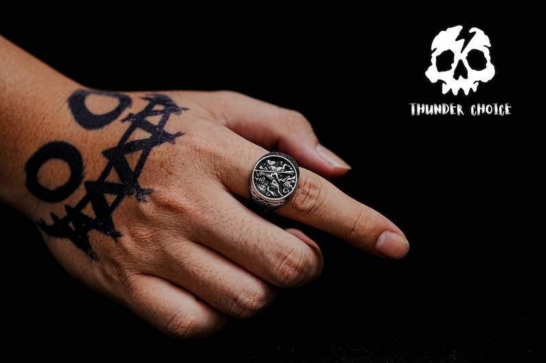 May include: A silver ring with a detailed design featuring a skull and crossbones. The ring is worn on the index finger of a hand with a black tattoo.