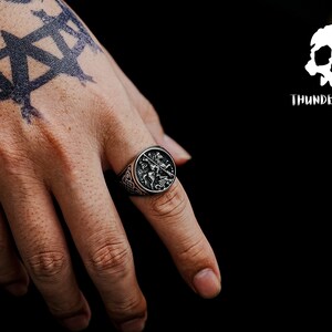 May include: A silver ring featuring a detailed design of a knight on horseback. The ring is worn on the index finger of a hand with a black tattoo.