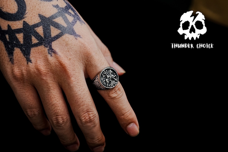 May include: A silver ring featuring a detailed design of a knight on horseback. The ring is worn on the index finger of a hand with a black tattoo.