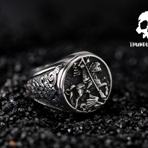May include: A silver ring featuring a detailed design of a knight on horseback. The ring has a textured band and a raised oval design depicting the knight.