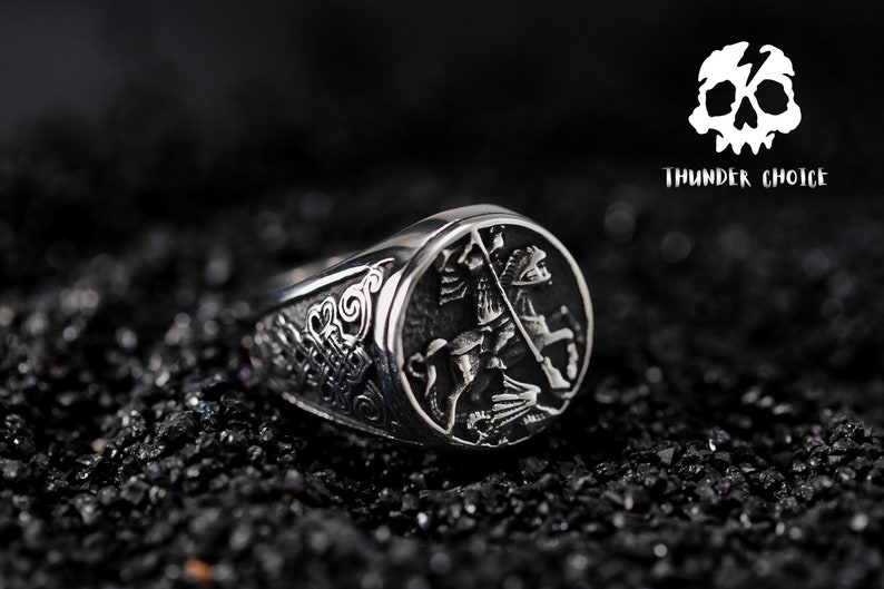 May include: A silver ring featuring a detailed design of a knight on horseback. The ring has a textured band and a raised oval design depicting the knight.