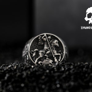 May include: A silver ring featuring a detailed design of a knight on horseback slaying a dragon. The ring is set against a black background.