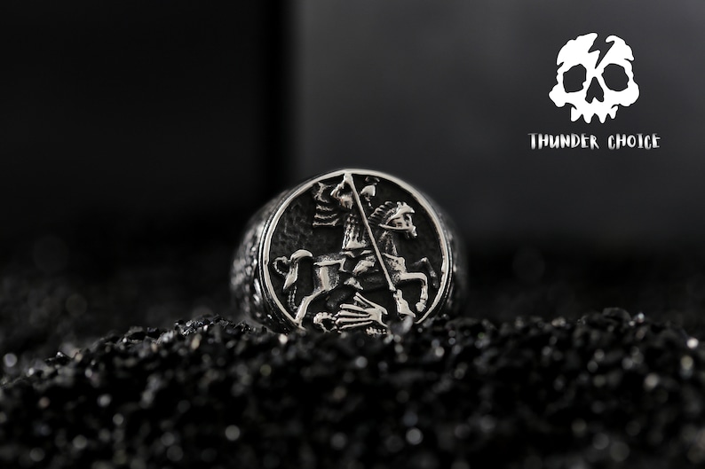 May include: A silver ring featuring a detailed design of a knight on horseback slaying a dragon. The ring is set against a black background.