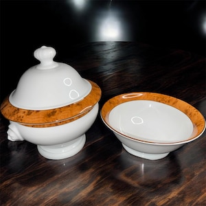 May include: A white porcelain serving bowl and a lidded dish with a decorative wood grain border. The dish has a rounded base and a knob on the lid. The bowl has a similar wood grain border.