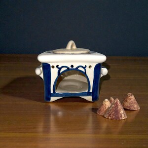 May include: A white and blue ceramic incense burner with a lid and handle, featuring a decorative blue design. The burner has a square shape with a rounded opening and is accompanied by several small, cone-shaped incense pieces.