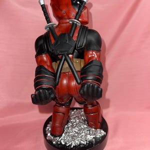 May include: A Deadpool figurine standing on a black circular base with silver debris. The character wears a red and black suit with two katanas crossed on the back, black gloves, and a utility belt. The figure is viewed from behind.