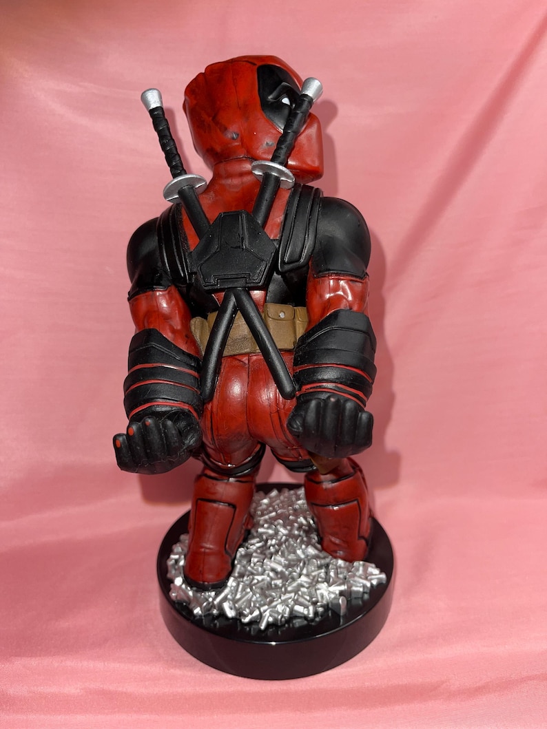 May include: A Deadpool figurine standing on a black circular base with silver debris. The character wears a red and black suit with two katanas crossed on the back, black gloves, and a utility belt. The figure is viewed from behind.