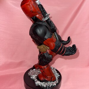 May include: A red and black character figurine standing on a circular black base with silver debris. The figure features a red mask, black torso, tan utility belt, and two swords strapped to the back. The hands are positioned forward as a holder.