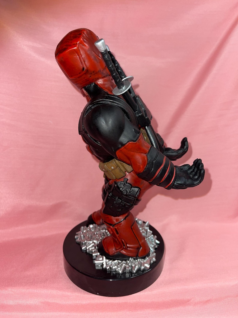 May include: A red and black character figurine standing on a circular black base with silver debris. The figure features a red mask, black torso, tan utility belt, and two swords strapped to the back. The hands are positioned forward as a holder.