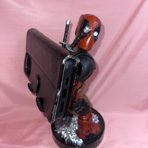 May include: A red and black Deadpool character figurine serving as a phone stand, holding a black rectangular smartphone case. The figure features two katana hilts protruding from the back and stands on a circular black base with silver accents.