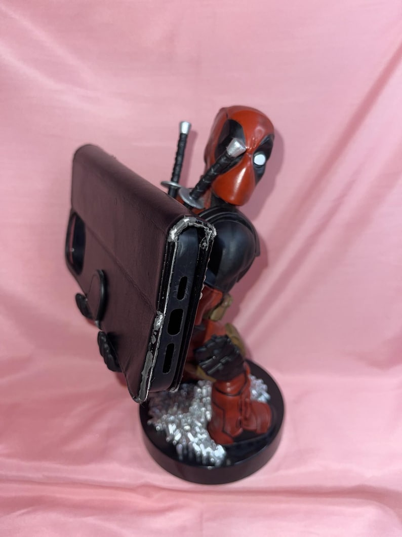May include: A red and black Deadpool character figurine serving as a phone stand, holding a black rectangular smartphone case. The figure features two katana hilts protruding from the back and stands on a circular black base with silver accents.