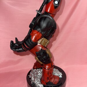 May include: A Deadpool figurine standing on a black circular base with silver debris. The character wears a red and black suit with a brown utility belt, holsters, and a sword hilt protruding from the back. The hands are positioned to hold an object.
