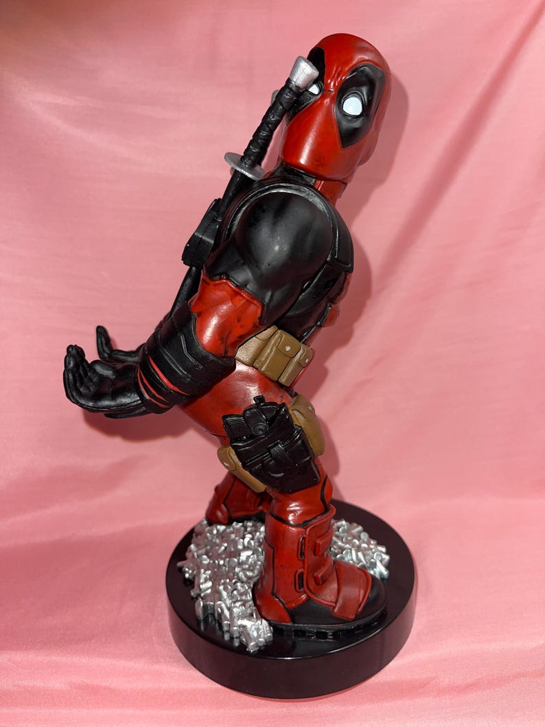 May include: A Deadpool figurine standing on a black circular base with silver debris. The character wears a red and black suit with a brown utility belt, holsters, and a sword hilt protruding from the back. The hands are positioned to hold an object.