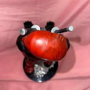 May include: A stylized figurine featuring a smooth, rounded red head with two silver-tipped sword hilts protruding from the back. The figure has a black body, stands on a circular black base, and includes a textured, light-colored debris detail.