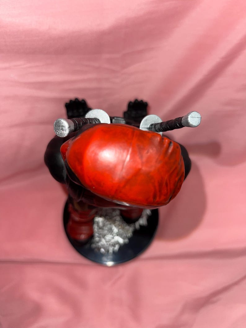 May include: A stylized figurine featuring a smooth, rounded red head with two silver-tipped sword hilts protruding from the back. The figure has a black body, stands on a circular black base, and includes a textured, light-colored debris detail.