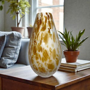 May include: A decorative glass vase with a speckled pattern in shades of amber and white. The vase has a teardrop shape and sits on a wooden table. A small potted plant and books are in the background.