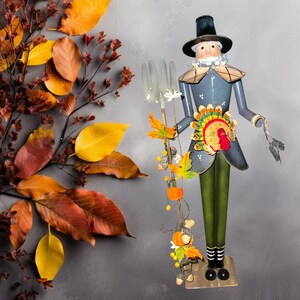May include: A metal figurine of a pilgrim holding a fork with a Thanksgiving-themed garland. The pilgrim is wearing a blue coat, green pants, and a black hat. The garland is decorated with pumpkins, acorns, and leaves.