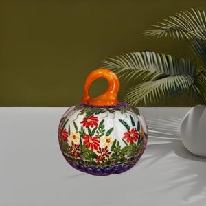 May include: A ceramic pumpkin-shaped decorative object featuring a glossy white base adorned with a vibrant floral pattern of red, yellow, and green botanical motifs. The piece includes a prominent, curved orange stem handle on top.