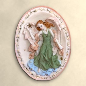 May include: An oval wall plaque featuring a relief sculpture of an angel with auburn hair, wearing a flowing green dress and large pink wings. The angel stands on blue clouds, holding a small star, surrounded by a border of tiny white flowers.