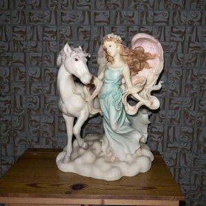May include: A detailed figurine of an angel with flowing hair and a light blue dress, standing beside a white horse. The angel has large, pink-tinged wings and a floral crown. The sculpture is set on a cloud-like base.