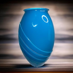 May include: A vibrant, glossy blue vase with a rounded, elongated shape. The vase features a series of curved, raised lines that wrap around its body. The design adds a unique texture to the smooth, solid color. The vase has a wide opening at the top.
