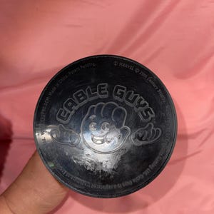 May include: A circular black plastic base for a Cable Guys figure featuring a central embossed cartoon hand icon and the text Cable Guys. The outer rim includes printed text referencing Marvel, 20th Century Studios, and Exquisite Gaming Ltd.