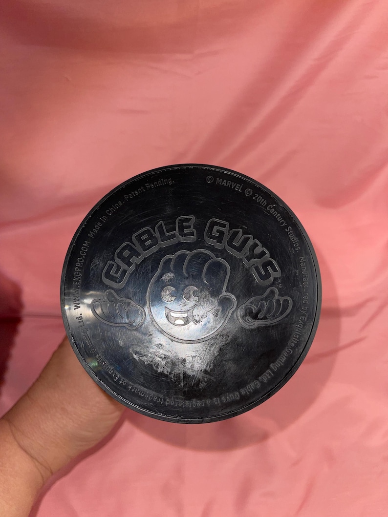 May include: A circular black plastic base for a Cable Guys figure featuring a central embossed cartoon hand icon and the text Cable Guys. The outer rim includes printed text referencing Marvel, 20th Century Studios, and Exquisite Gaming Ltd.