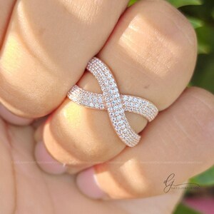 May include: A rose gold crossover ring featuring a crisscross design encrusted with numerous small, clear, sparkling pav&eacute;-set stones. The band curves into an X-shape, creating a continuous, shimmering surface across the top of the jewelry piece.
