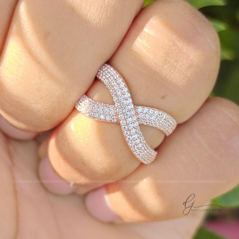 May include: A rose gold crossover ring featuring a crisscross design encrusted with numerous small, clear, sparkling pav&eacute;-set stones. The band curves into an X-shape, creating a continuous, shimmering surface across the top of the jewelry piece.