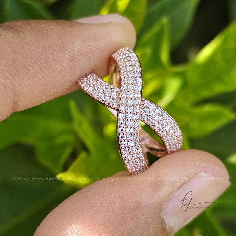 May include: A rose gold crossover ring featuring a pav&eacute; setting with numerous small, clear, round-cut stones covering the entire surface of the band. The ring has an X-shaped design where the two bands intersect, creating a continuous sparkling effect.