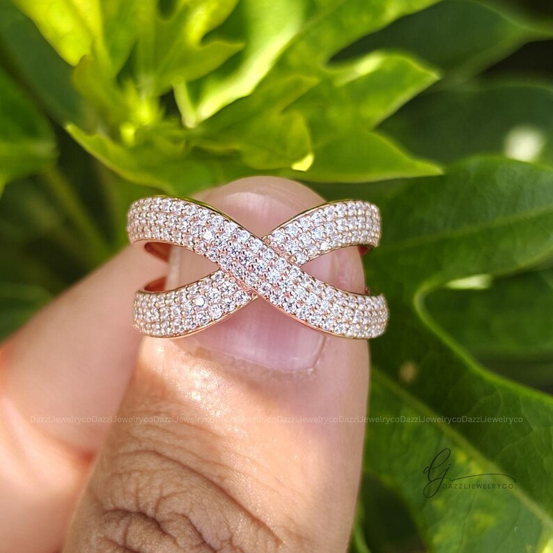 May include: A rose gold crossover ring featuring two intersecting bands completely encrusted with small, clear, sparkling pav&eacute;-set stones. The design creates an X-shaped silhouette with a polished metal finish visible along the edges of the bands.