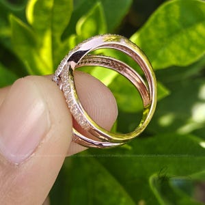 May include: A rose gold ring featuring a crossover design with two bands that intersect. The surface of the bands is encrusted with numerous small, clear, sparkling stones. The metal has a polished, reflective finish throughout the entire structure.