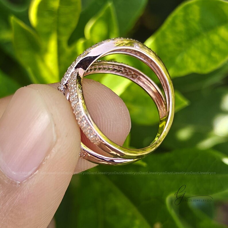 May include: A rose gold ring featuring a crossover design with two bands that intersect. The surface of the bands is encrusted with numerous small, clear, sparkling stones. The metal has a polished, reflective finish throughout the entire structure.