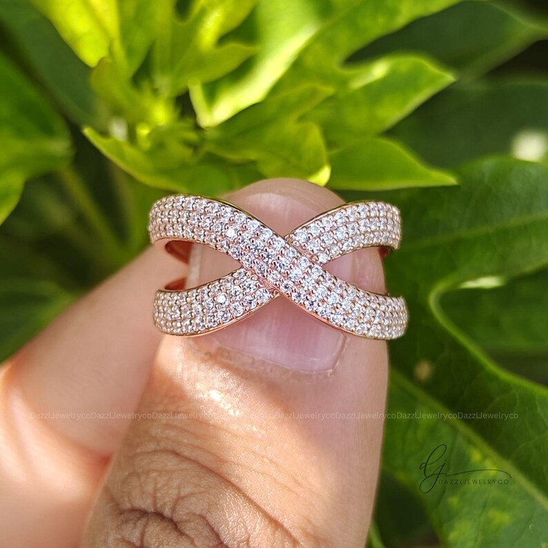 May include: A rose gold crossover ring featuring an X-shaped design completely encrusted with small, clear, sparkling pav&eacute;-set stones. The band splits into two intersecting paths that form a continuous, symmetrical, and wide metallic structure.