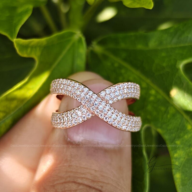 May include: A rose gold-toned crossover ring featuring a wide band design that forms an X shape. The entire surface of the ring is encrusted with numerous small, clear, sparkling stones set closely together in a pav&eacute; style across the metal.